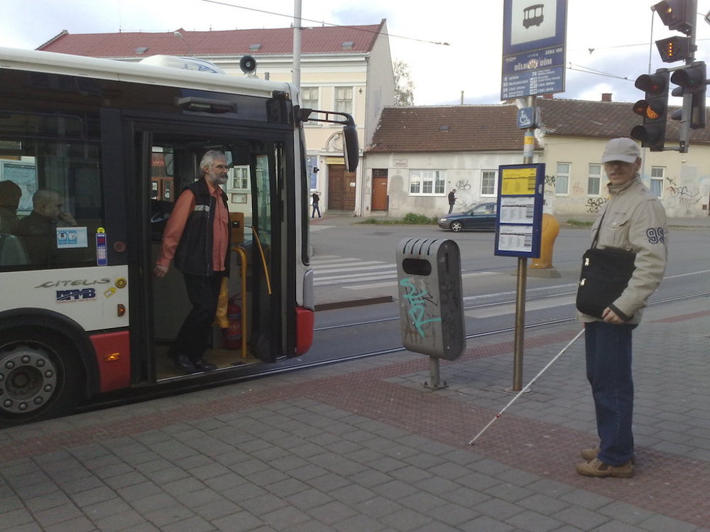Na obrázku je nevidomý cestující u označníku zastávky. Před ním otevřené dveře trolejbusu. Nevidomý nijak nereaguje, protože neví, že trolejbus už přijel.